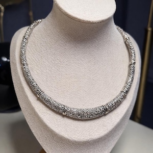 May include: A silver-colored necklace with an intricate, detailed pattern. The necklace is displayed on a cream-colored bust, showcasing its design. The necklace appears to be made of metal and has a textured surface.