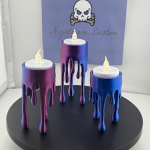 May include: Three tealight candle holders with a dripping wax design. The candle holders are in shades of purple and blue. The candle holders are on a black circular base. The text "Nightmare Custom" is visible in the background.