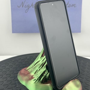 Puede incluir: Un soporte para teléfono impreso en 3D de color verde y rojo con una funda de teléfono negra en posición vertical. El soporte para teléfono tiene forma de montaña con una superficie texturizada.