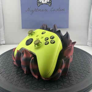 May include: A lime green Xbox controller sits in a black and red 3D printed stand. The stand is shaped like a flame and has a textured surface. The stand is on a black turntable. The text "Nightmare Custom" is written on a purple background behind the controller.