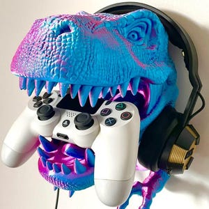 Peut inclure: Support mural en forme de tête de dinosaure bleue et rose, tenant une manette de jeu blanche. Un casque noir est monté sur le côté. Conçu pour ranger des accessoires de jeu.