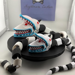 Pode incluir: Duas esculturas de serpentes com cabeça de tubarão com detalhes brancos, vermelhos e azuis. As serpentes são pretas e brancas, com uma cauda pontiaguda. Uma placa no fundo diz "Nightmare Custom" com um logotipo de caveira e ossos cruzados.