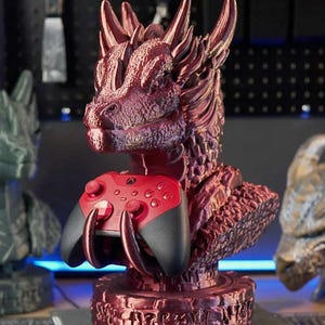 Puede incluir: Un busto de dragón burdeos sosteniendo un mando de Xbox rojo y negro. El dragón tiene escamas y cuernos detallados, y el mando tiene botones y joysticks. La base del busto tiene un diseño grabado. Altura aproximada: 20 cm.