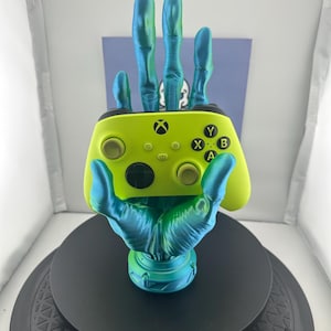 May include: A bright green Xbox controller is held by a teal blue 3D printed hand. The hand is on a black circular base.