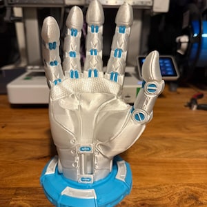Könnte beinhalten: Eine weiße, 3D-gedruckte Roboterhand mit blauen Akzenten, die mit ausgestreckten Fingern posiert, auf einem blauen Sockel montiert.