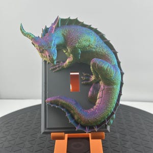 May include: A 3D printed iridescent dragon light switch cover. The dragon is curled around the switch plate and has a green, blue, and purple iridescent finish.
