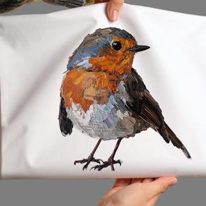 May include: A white rectangular fabric item featuring a textured, impasto-style painting of a European robin. The bird has a bright orange breast, a grey-brown head and back, and dark wings, with visible brushstrokes and small printed text details.