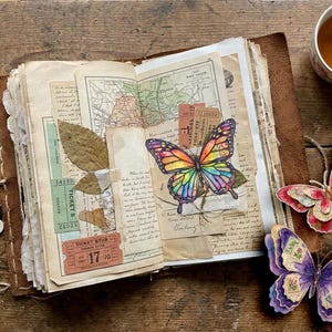 Puede incluir: Un diario abierto encuadernado en cuero con p&aacute;ginas de collage que incluyen mapas antiguos, talones de boletos, hojas secas y una ilustraci&oacute;n de mariposa colorida. Dos recortes de mariposas de papel superpuestas descansan cerca, junto a llaves.