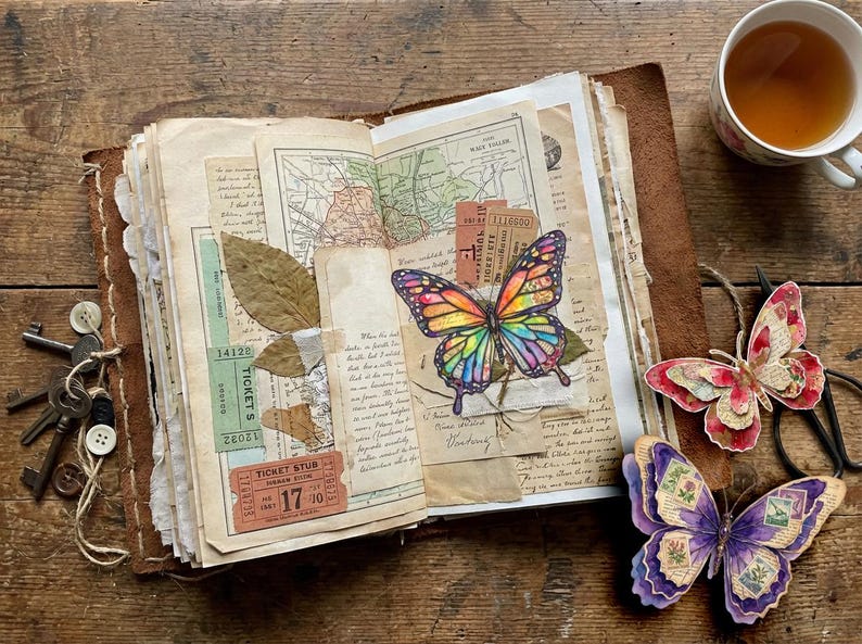 Puede incluir: Un diario abierto encuadernado en cuero con p&aacute;ginas de collage que incluyen mapas antiguos, talones de boletos, hojas secas y una ilustraci&oacute;n de mariposa colorida. Dos recortes de mariposas de papel superpuestas descansan cerca, junto a llaves.