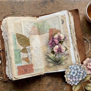 Op de afbeelding: Een open dagboek met een bruine leren kaft, versierd met vintage ephemera zoals een landkaart, toegangskaartjes en gedroogde bladeren. De pagina's zijn gedecoreerd met illustraties van paarse orchidee&euml;n, blauwe hortensia's en een roze pioenroos.