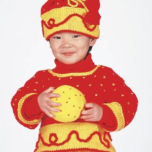 May include: A child wearing a red and yellow knitted sweater and hat, holding a yellow ball. The sweater has a yellow trim and a yellow and red pattern. The hat has a yellow and red pattern and a pom-pom on top.