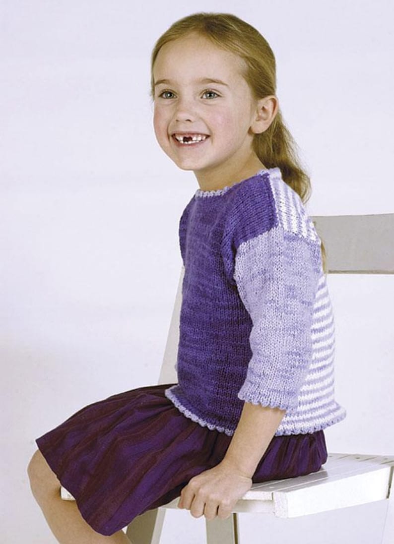 Puede incluir: Un su&eacute;ter morado tejido para ni&ntilde;os con un dise&ntilde;o de bloques de color. Las mangas son de color lila claro y a rayas blancas. La ni&ntilde;a lleva una falda morada oscura y est&aacute; sentada en una silla blanca.