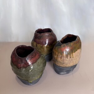 May include: Three small, handcrafted ceramic vases. Each vase has a unique shape and colour palette, with a dark interior and a mix of green, brown, and red tones on the exterior. The bases are a dark grey.