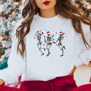 May include: White sweatshirt featuring a graphic of three dancing skeletons wearing Santa hats and Christmas lights. The skeletons are in various poses, with snowflakes around them. The sweatshirt is a festive holiday design.
