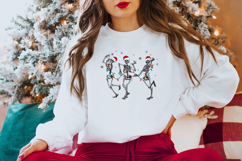May include: White sweatshirt featuring a graphic of three dancing skeletons wearing Santa hats and Christmas lights. The skeletons are in various poses, with snowflakes around them. The sweatshirt is a festive holiday design.