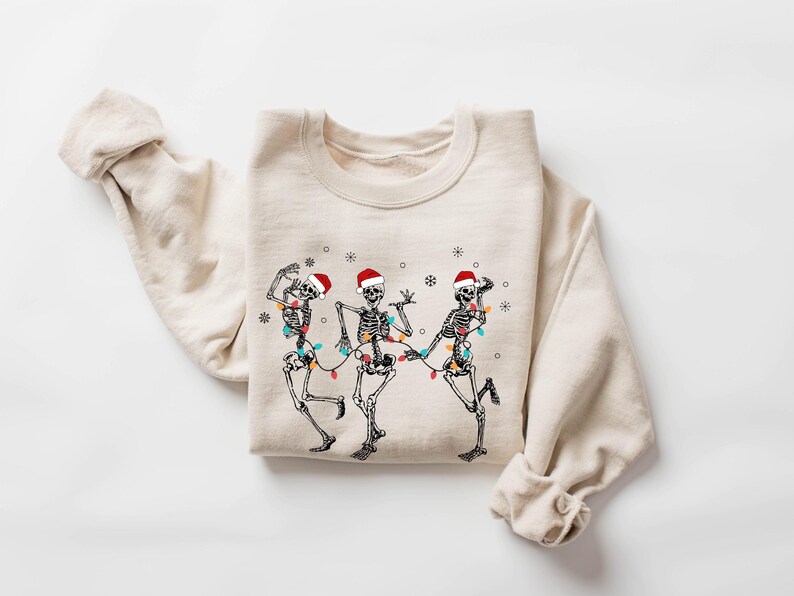 May include: Beige sweatshirt featuring a graphic of three dancing skeletons wearing Santa hats and strung with Christmas lights. The design is in black and white with red and multicolored accents, perfect for the holiday season.