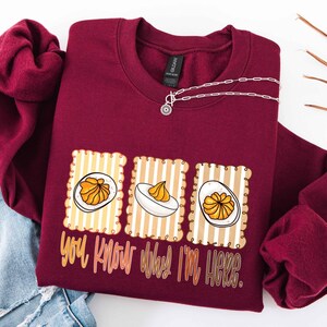 May include: Burgundy sweatshirt with a graphic of deviled eggs on a striped background. The text "You know why I'm here" is below the graphic. A silver necklace with a small pendant is on the sweatshirt.