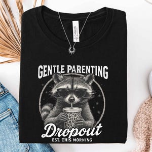 May include: Black t-shirt featuring a raccoon wearing sunglasses and drinking from a leopard print cup. The text reads "GENTLE PARENTING" above the raccoon and "Dropout EST. THIS MORNING" below.