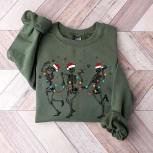 May include: An olive green sweatshirt featuring a graphic of three dancing skeletons wearing Santa hats and strung with Christmas lights. The design is printed on the front of the long-sleeved crew neck top. The sweatshirt is folded on a light wood-grain surface.