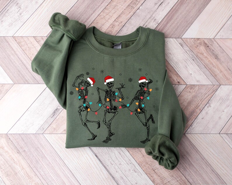 May include: An olive green sweatshirt featuring a graphic of three dancing skeletons wearing Santa hats and strung with Christmas lights. The design is printed on the front of the long-sleeved crew neck top. The sweatshirt is folded on a light wood-grain surface.