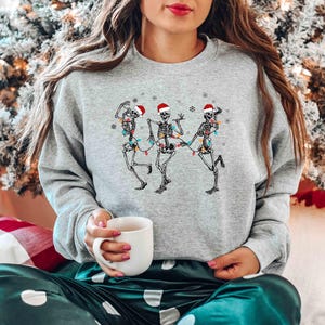 May include: A light gray sweatshirt featuring a graphic of three dancing skeletons wearing Santa hats and strung with Christmas lights. The skeletons are in a festive pose, perfect for the holiday season.