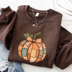 May include: A brown sweatshirt featuring a patchwork pumpkin design with a gingham bow. The pumpkin is composed of various fabric patterns in orange, green, and tan. A silver necklace with a small pendant rests on the sweatshirt.