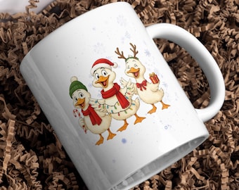 Op de afbeelding: Witte keramische mok met een feestelijk ontwerp van drie cartoon eenden gekleed voor Kerstmis. Eén eend draagt een kerstmuts, een andere heeft rendiergeweien en de derde houdt een zuurstok vast. De eenden zijn versierd met sjaals en lichtjes, met sneeuwvlokken op de achtergrond.