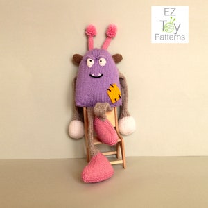 Könnte beinhalten: Ein lila gestricktes Monster-Spielzeug mit braunen Armen und rosa Beinen, das auf einem Holzstuhl sitzt. Das Monster hat einen gelben Fleck auf seinem Körper und ein lächelndes Gesicht. Das Spielzeug wird von EZ Toy Patterns hergestellt.