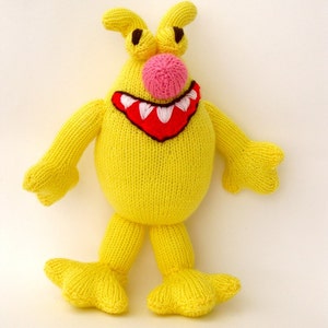 May include: A yellow knitted toy monster with a big smile, red mouth, white teeth, and a pink nose. The monster has two large eyes and two small arms.
