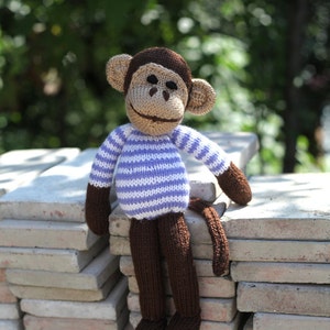 May include: A knitted monkey toy with brown fur, a purple and white striped shirt, and a brown tail. The monkey is sitting on a stack of gray concrete blocks.
