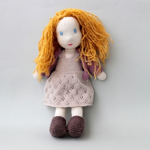 May include: Handmade knitted doll with a white head, blue eyes, and red lips. The doll has long, orange yarn hair, a light purple dress, and a matching jacket. The doll's feet are brown, and it is standing on a light gray surface.