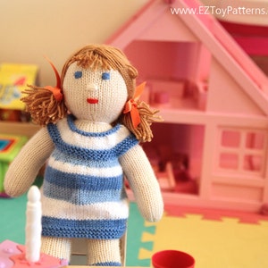Peut inclure: Une poupée en tricot avec des cheveux bruns, une robe rayée bleu et blanc et des nœuds orange. La poupée est assise devant une maison de jeu rose.