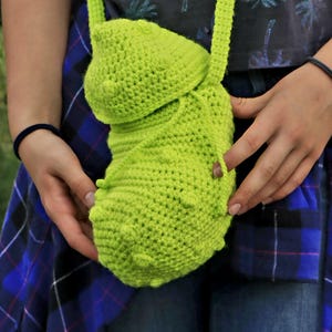 May include: A lime green crochet bag with a long strap and a rounded shape. The bag has a textured surface with small, raised bumps and a wooden button closure. The bag is held by a person wearing a blue plaid shirt.