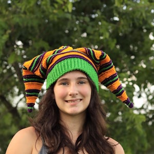 May include: A crochet jester hat featuring a bright green ribbed brim and two long, pointed horns extending from the sides. The hat is decorated with horizontal stripes in alternating colors of vibrant orange, deep purple, and black yarn throughout.