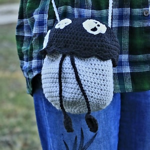 May include: A gray crocheted bag with black accents and white skull details. The bag has a long strap and is worn over the shoulder.