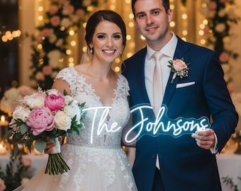 May include: A newly married couple smiles for a photo. The bride holds a bouquet of pink and white flowers, and wears a white lace wedding dress. The groom wears a navy blue suit and holds a neon sign reading "The Johnsons".