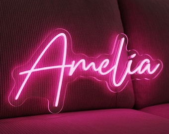 Könnte beinhalten: Ein Neon-Schild mit dem Namen "Amelia" in kursiver Schrift, leuchtend in hellem Pink. Das Schild ist vor einem dunkelrosa, strukturierten Hintergrund platziert, wahrscheinlich einem Sofa oder Kissen.