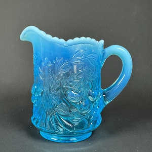 May include: A vintage, light blue glass jug with a detailed floral design. The jug has a scalloped rim, a curved handle, and a round base. The glass has a slight opalescent quality.