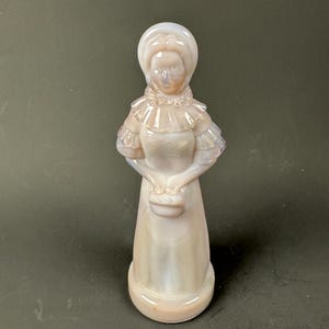 May include: A vintage figurine of a woman in a bonnet and dress, holding a small purse. The figurine is made of opalescent glass, with a soft pink and white color scheme. The woman's dress features ruffled details. The figurine is standing on a round base.