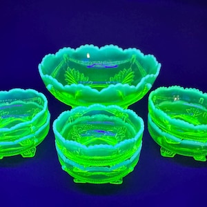 May include: A collection of green glass bowls and dishes, including a large serving bowl and several smaller bowls. The glass has a scalloped edge and a decorative pattern. The bowls are illuminated with a bright green glow against a dark blue background.