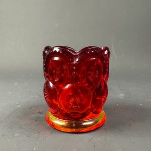 Op de afbeelding: Een robijnrode glazen uilvormige container met een goudkleurige basis. De container heeft een gedetailleerd ontwerp met zichtbare ogen en veren. De uil is een decoratief item, waarschijnlijk voor het bewaren van kleine items.