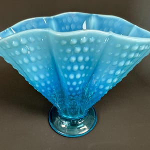 May include: A light blue hobnail glass vase with a ruffled rim. The vase has a wide, fan-like shape and sits on a round base. The glass has a textured surface with small, raised dots. The color is a vibrant, translucent blue.