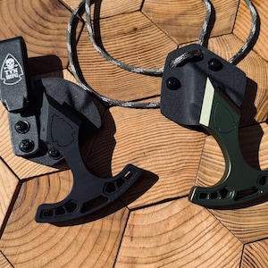 Könnte beinhalten: Zwei schwarze und grüne Halsmesser mit Scheiden und Paracord-Lanyards. Eines ist schwarz, das andere grün mit einem weißen Streifen. Auf den Scheiden steht der Text "TYN MFG" und ein Totenkopf-Logo.