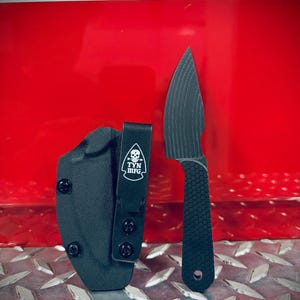 Puede incluir: Un cuchillo negro de hoja fija con mango texturizado y funda a juego. La hoja del cuchillo tiene un patrón estriado único. La funda presenta el texto "TYN MFG" con un logotipo de calavera. El fondo es rojo y de metal con diseño de diamante.