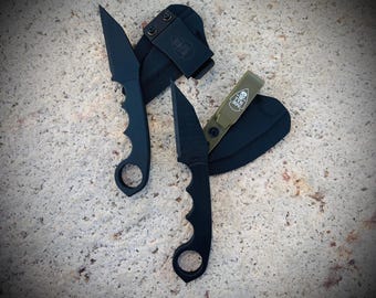 Può includere: Due coltelli tattici neri con manico ad anello e lama a punta sono esposti. Ogni coltello è abbinato a un fodero nero. Un fodero ha una clip da cintura, l'altro è verde oliva con un logo a teschio.