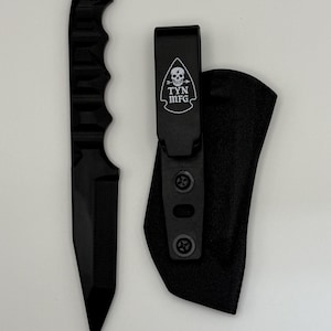 May include: A black fixed-blade knife with a textured handle and a sheath. The sheath has a clip with the text "TYN MFG" and a skull logo. The knife is designed for utility and outdoor use.