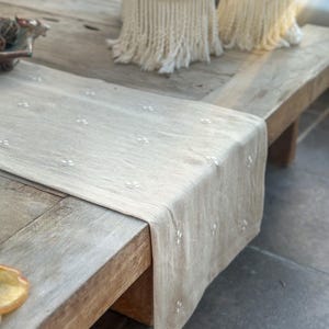 May include: A beige linen table runner with embroidered floral details, draped over a rustic wooden table. Two macrame candle holders sit on the table. The runner adds a touch of elegance to the natural wood.