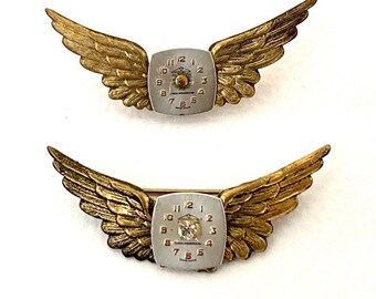 May include: Two gold-tone winged brooches with a silver clock face in the center. The top brooch has a plain clock face, while the bottom brooch has a small diamond in the center of the clock face.