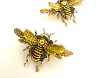 May include: Two gold bee brooches with yellow and black striped bodies, yellow wings, and a yellow flower detail on the head. The brooches are on a white background.
