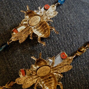 May include: Gold bee charm necklace with a blue and silver chain. The bee charm has the text "CARA STIMMEL Ltd." on it.